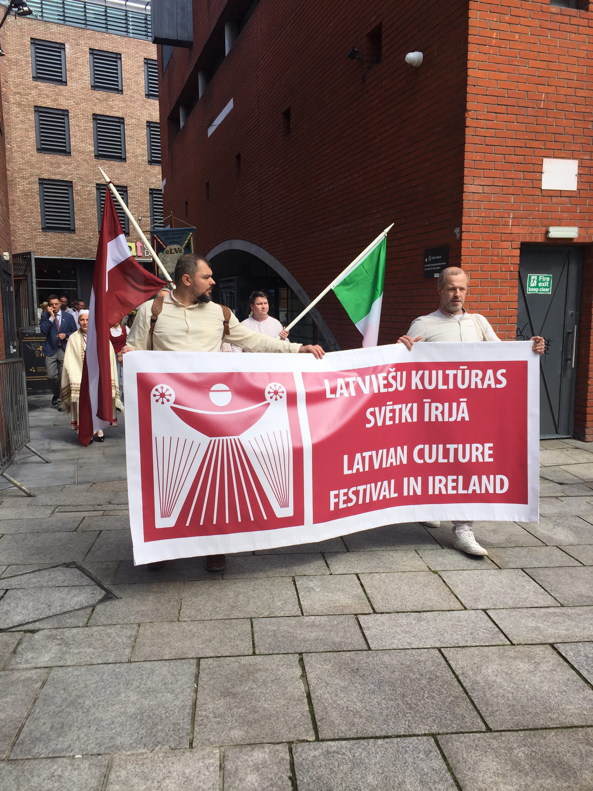 Cilvēki nes plakātu - Latviešu kultūras svētki Īrijā. Fonā diviem cilvēkiem redzami citi gājiena dlaībnieki, Īrijas un Latvijas karogi.