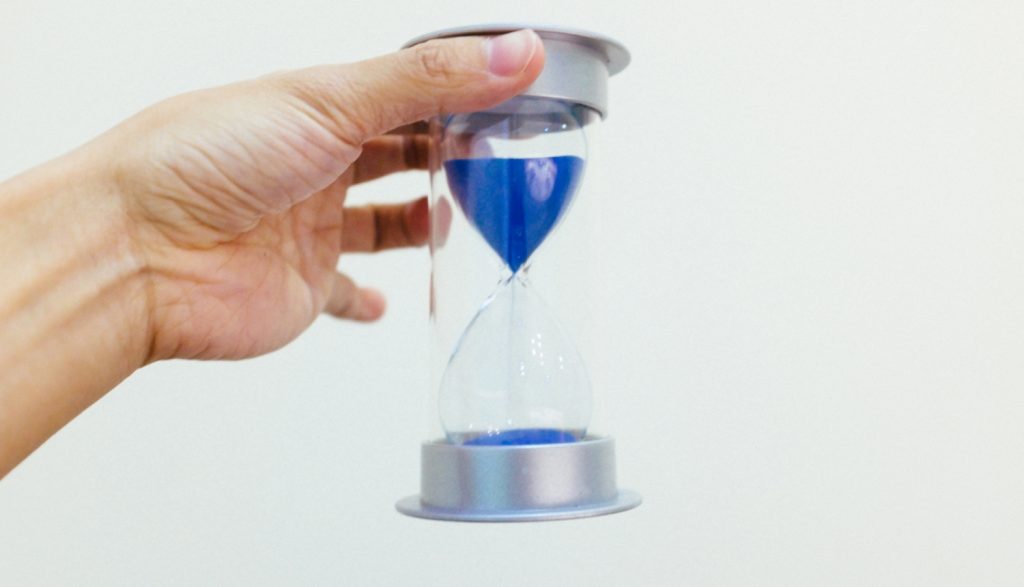 Ilustratīvs attēls - roka ar smilšu pulksteni, kurā ir zilas krāsas smiltis.