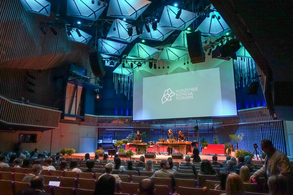 Kurzemes biznesa foruma norises zāles foto - redzama skatuve zili zaļos toņos ar lielu ekrānu un zālē sēdoši cilvēki - aptuveni 30 cilvēki.
