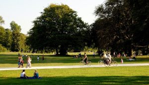 Ilustratīvs foto, parks vasarā ar kokiem, zaļu zāli, pa baltu celiņu pastaigājas vairāki vasarīgi tērpti cilvēki.